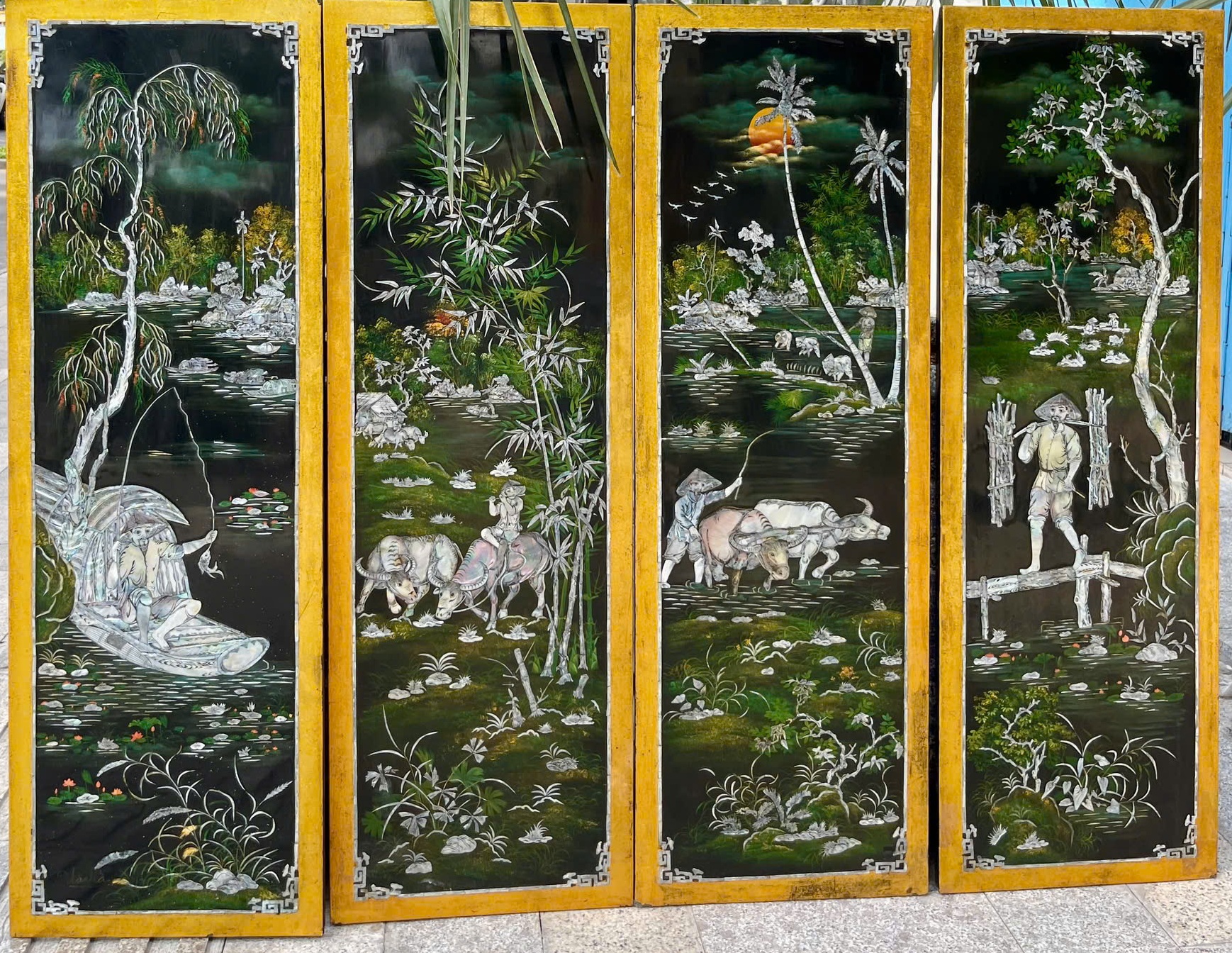 Ngư tiều canh mục — Four slender panels bring rural life into view: fishing boats, bamboo, buffalo and figures crossing waterways. Mother-of-pearl inlay catches the light against the dark lacquer ground.